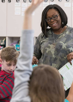 A teacher in a classroom smiling while students raise their hands.